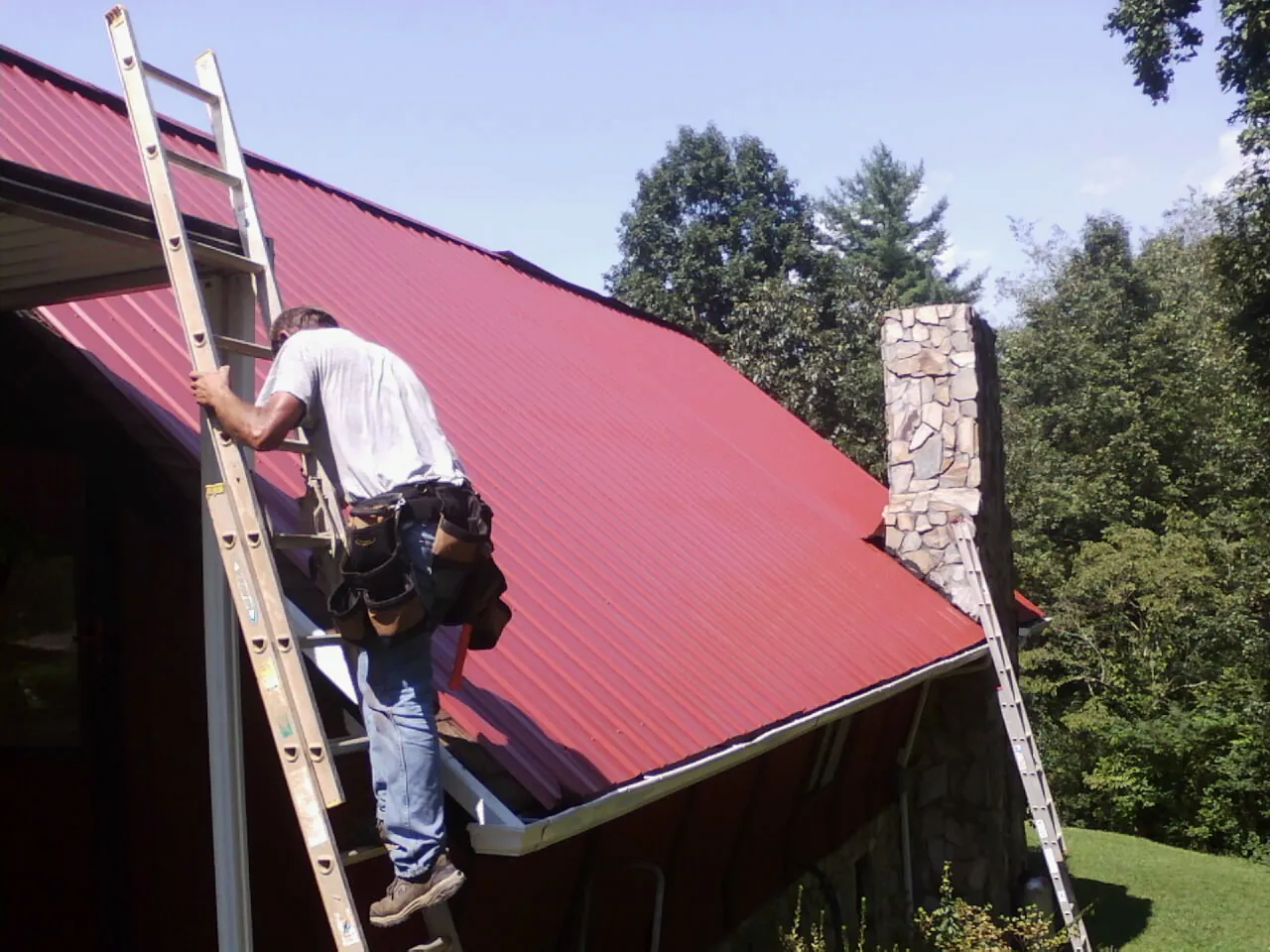 Roofer on a ladder beside a red metal roof and stone chimney — Soffit & Fascia Repair by Roofing Brothers in Lewiston Woodville